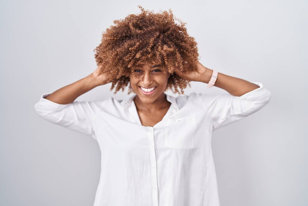 Молода іспаномовна жінка з кучерявим волоссям, що стоїть над білим тлом, посміхаючись тягнути вуха пальцями, смішний жест. проблема прослуховування  - Фото, зображення