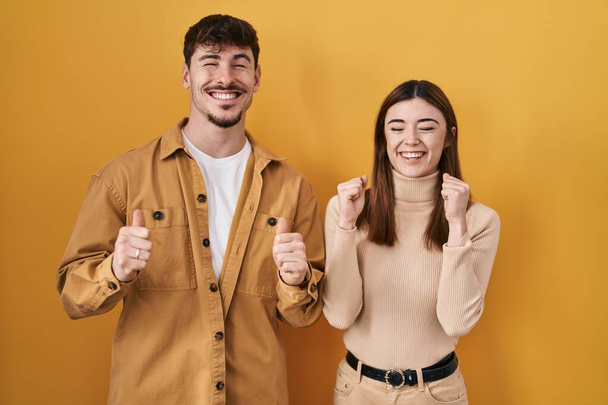 Mladý hispánský pár stojící nad žlutým pozadím nadšený úspěchem se zdviženýma rukama a zavřenýma očima oslavující vítězství s úsměvem. koncept vítěze.  - Fotografie, Obrázek