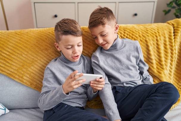 Uroczy chłopcy oglądanie wideo na smartfonie siedzi na kanapie w domu - Zdjęcie, obraz