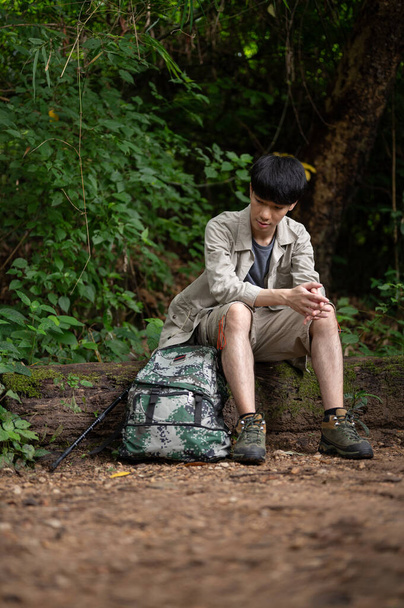 An exhausted young Asian male hiker sits on a stone, resting and taking a break during his hiking trip in the forest alone. - Фото, зображення