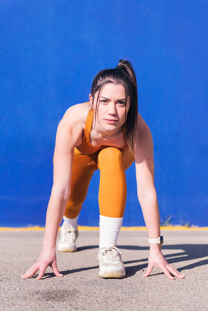 vertical portrait of a focused sportswoman in position to start a race with orange sportswear in a blue background, concept of sport and active lifestyle, copy space for text - Фото, изображение
