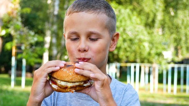 Roztomilý devítiletý chlapec si v létě užívá hamburger v městském parku. Koncept dětské výživy, rychlého občerstvení na ulici. Ulice rychlé občerstvení - Fotografie, Obrázek