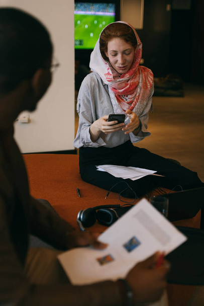 Linda gengibre muçulmano empregado verificando seu telefone ao ter uma conversa com seu colega masculino preto - Foto, Imagem