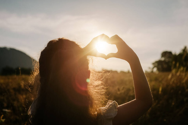 Back view of little girl child making heart with hands during calm sunset or sunrise at warm summer day. Sunbeams Sunlight. Happy Valentines day, Mothers Day, love nature or life happiness concept - Foto, imagen