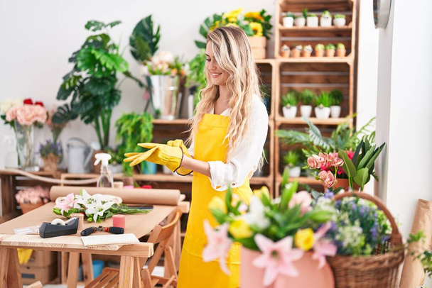 Nuori blondi nainen kukkakauppa hymyilee luottavainen yllään käsineet kukkakauppa - Valokuva, kuva