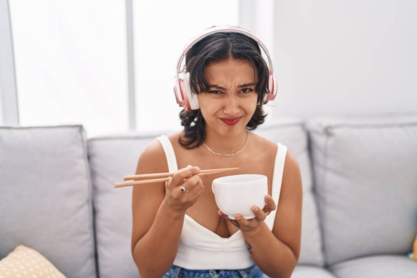Mujer hispana joven comiendo comida asiática usando palillos de expresión despistada y confusa. concepto de duda.  - Foto, imagen