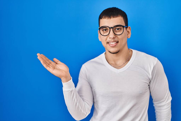 Jeune homme arabe portant une chemise blanche décontractée et des lunettes souriantes et joyeuses présentant et pointant la paume de la main en regardant la caméra.  - Photo, image