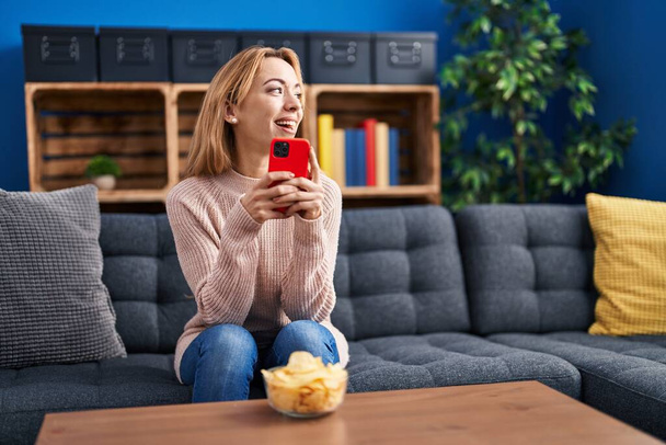 Jeune femme utilisant smartphone manger des pommes de terre chips à la maison - Photo, image
