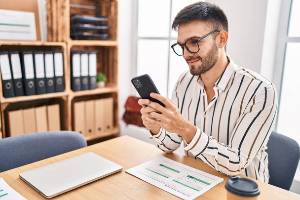 Молодий іспаномовний чоловік бізнесмен посміхається впевнено використовуючи смартфон в офісі
 - Фото, зображення