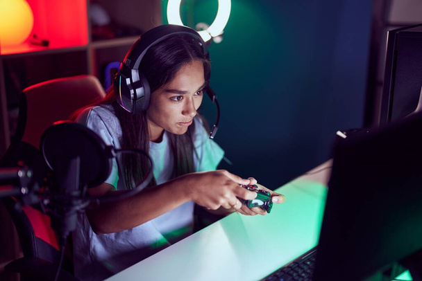 Young beautiful hispanic woman streamer playing video game using joystick at gaming room - Photo, image