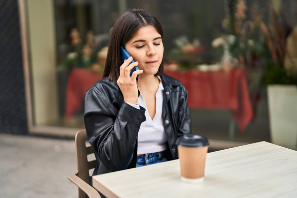 Молодая красивая латиноамериканка разговаривает на смартфоне, сидя на столе на террасе кафе - Фото, изображение