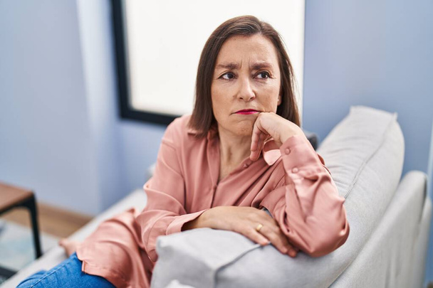 Kobieta w średnim wieku ze smutnym wyrazem twarzy siedzi na kanapie w domu - Zdjęcie, obraz