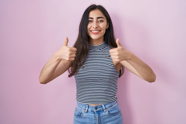 Young teenager girl wearing casual striped t shirt approving doing positive gesture with hand, thumbs up smiling and happy for success. winner gesture.  - Photo, Image