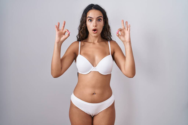 Mujer hispana joven vistiendo lencería blanca que se ve sorprendida y sorprendida haciendo símbolo de aprobación aceptable con los dedos. expresión loca  - Foto, imagen
