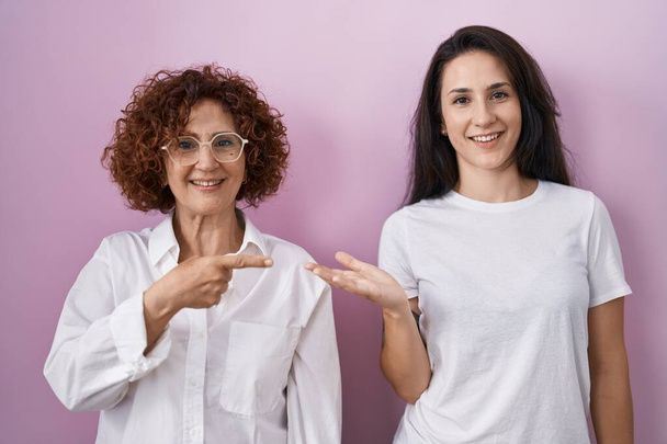 Hispánská matka a dcera na sobě ležérní bílé tričko přes růžové pozadí ohromený a usmívá se do kamery při prezentaci s rukou a ukazuje prstem.  - Fotografie, Obrázek