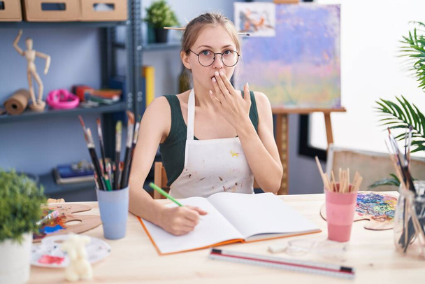 Mladá běloška sedící v ateliéru umění maluje rukou na zápisník, zakrývající ústa, šokovaná a vystrašená z omylu. překvapený výraz  - Fotografie, Obrázek