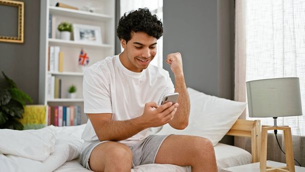 Young latin man using smartphone sitting on bed celebrating at bedroom - Фото, изображение