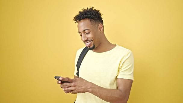 Afro-Amerikaanse man draagt rugzak met behulp van smartphone over geïsoleerde gele achtergrond - Foto, afbeelding