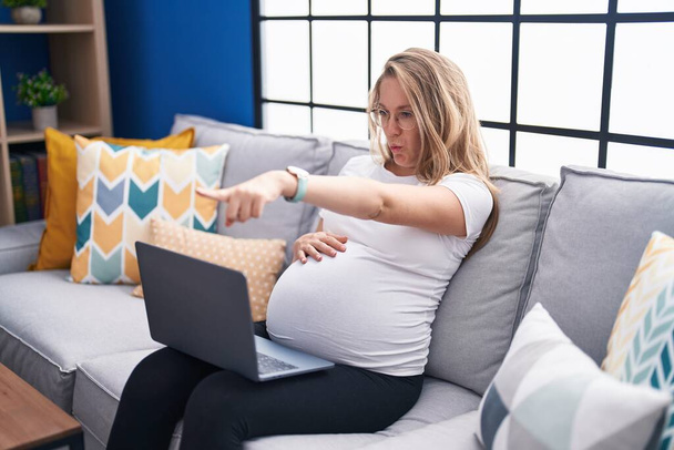 Νεαρή έγκυος γυναίκα κάθεται στον καναπέ στο σπίτι χρησιμοποιώντας το laptop δείχνοντας με το δάχτυλο στην κάμερα και σε σας, αυτοπεποίθηση χειρονομία αναζητούν σοβαρή  - Φωτογραφία, εικόνα