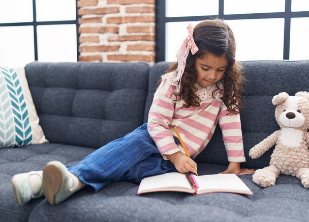 Adorable hispanic girl drawing on notebook sitting on sofa at home - 写真・画像