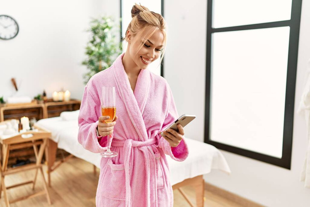Jovem caucasiana vestindo roupão de banho bebendo champanhe usando smartphone no salão de beleza - Foto, Imagem