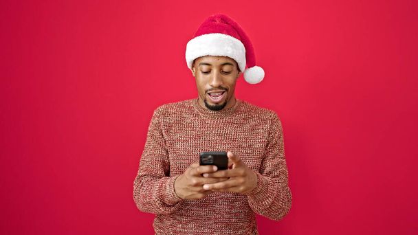 Afrykański Amerykanin nosi świąteczny kapelusz za pomocą smartfona uśmiechając się na odizolowanym czerwonym tle - Zdjęcie, obraz