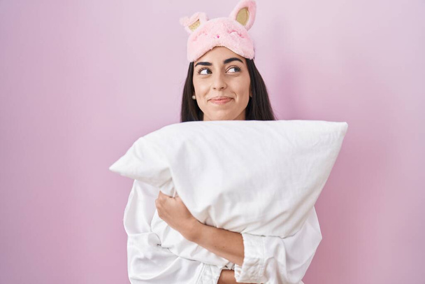 Young brunette woman wearing sleep mask and pajama hugging pillow smiling looking to the side and staring away thinking.  - Photo, Image