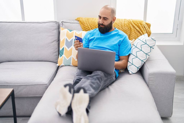 Νεαρός φαλακρός άντρας που χρησιμοποιεί φορητό υπολογιστή και πιστωτική κάρτα κάθεται στον καναπέ στο σπίτι - Φωτογραφία, εικόνα