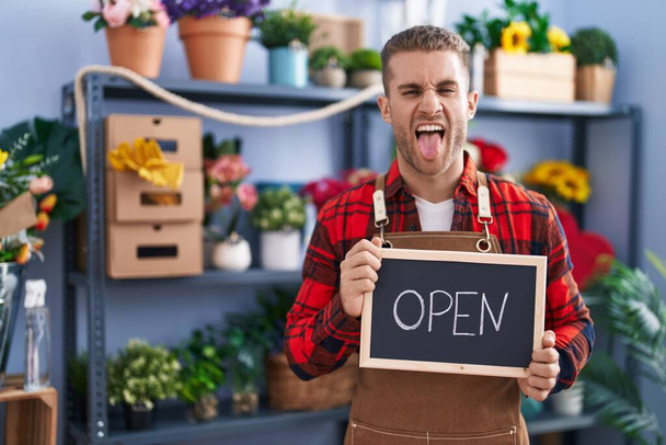 Young caucasian man working at florist holding open sign sticking tongue out happy with funny expression.  - Photo, Image