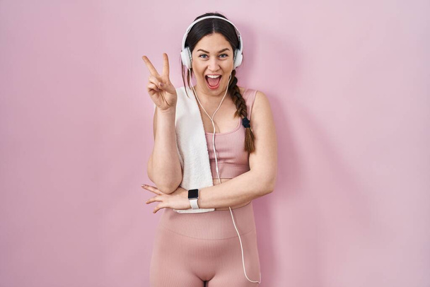 Young brunette woman wearing sportswear and headphones smiling with happy face winking at the camera doing victory sign. number two.  - Foto, Imagem