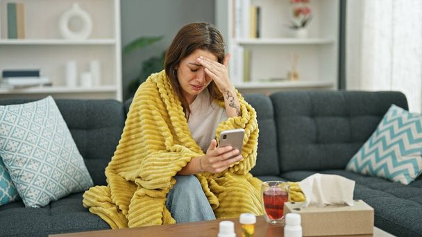 Mladá krásná hispánská žena sedí na pohovce nemocný s online lékařskou konzultaci na smartphone doma - Fotografie, Obrázek