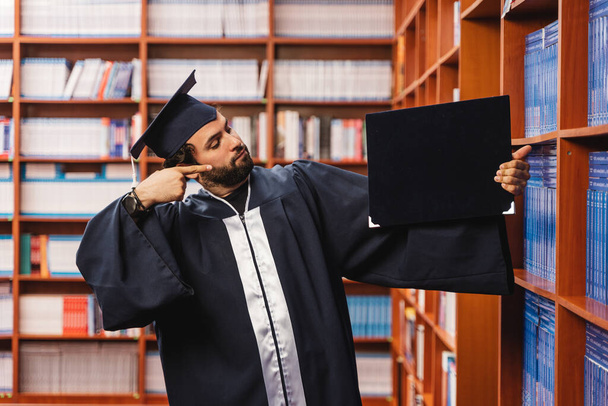 Young and happy graduate, wearing a cap and gown while pointing towards his bachelor's degree in a library - Photo, Image
