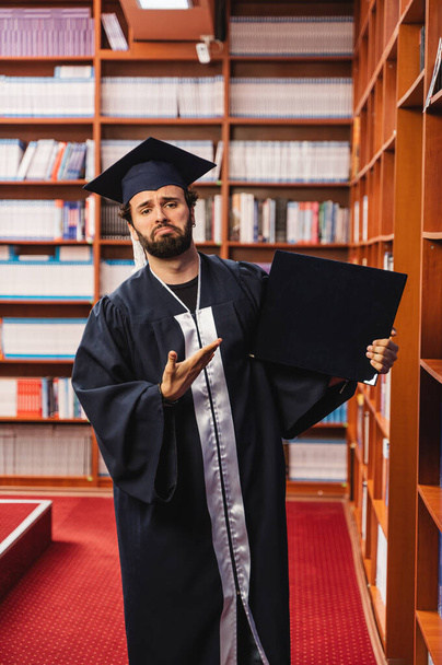 Young graduate wearing his cap and gown looking sad while holding his university degree - Photo, Image
