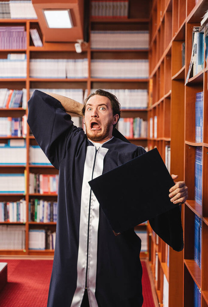 Giovane laureato indossa un abito scioccato mentre tiene la sua laurea in una biblioteca - Foto, immagini