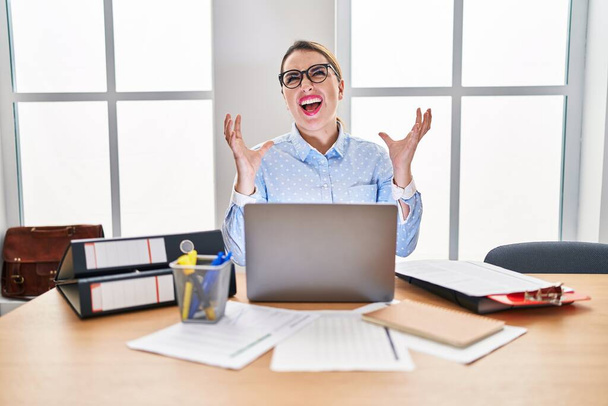 Young hispanic woman working at the office wearing glasses crazy and mad shouting and yelling with aggressive expression and arms raised. frustration concept.  - Foto, imagen