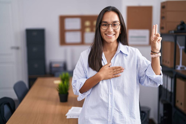 Mujer hispana joven en la oficina sonriendo jurando con la mano en el pecho y los dedos hacia arriba, haciendo un juramento de lealtad promesa  - Foto, imagen
