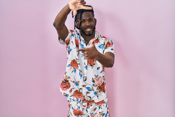 Uomo africano con dreadlocks indossa camicia estiva su sfondo rosa sorridente rendendo cornice con le mani e le dita con il viso felice. creatività e concetto di fotografia.  - Foto, immagini