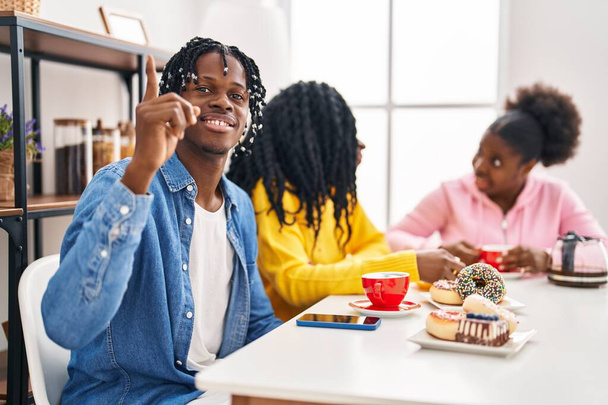 Gruppo di tre giovani neri seduti su un tavolo a bere caffè sorpresi da un'idea o da una domanda che punta il dito con la faccia felice, numero uno  - Foto, immagini