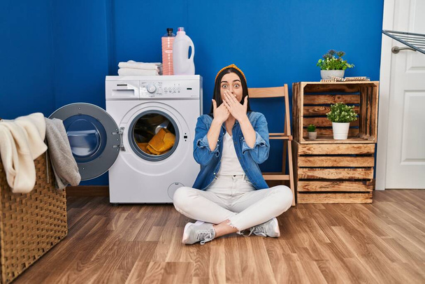 Іспаномовна жінка, яка займається пранням, сидячи на підлозі, шокувала, покриваючи рот руками за помилку. секретна концепція.  - Фото, зображення