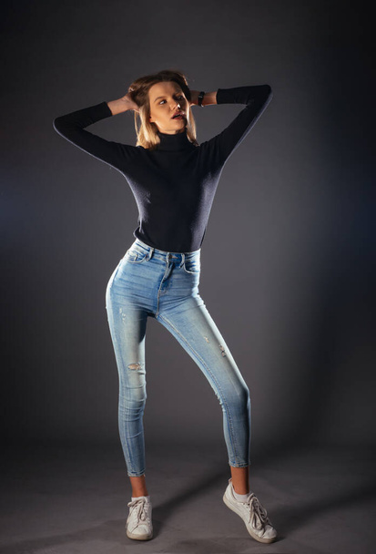 Foto a figura intera di una bella modella in posa su sfondo scuro in uno studio con camicetta nera, jeans e scarpe da ginnastica bianche - Foto, immagini