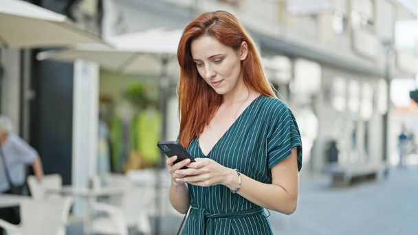 Giovane donna rossa utilizzando smartphone con espressione seria sulla terrazza della caffetteria - Foto, immagini