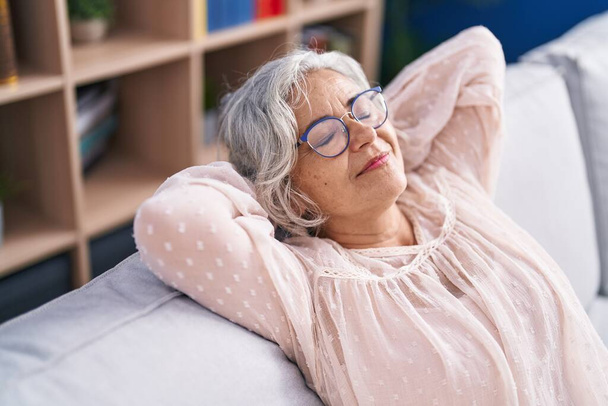 Femme aux cheveux gris d'âge moyen détendue avec les mains sur la tête assise sur le canapé à la maison - Photo, image