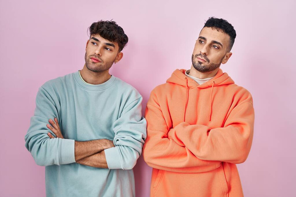 Young hispanic gay couple standing over pink background looking to the side with arms crossed convinced and confident  - 写真・画像