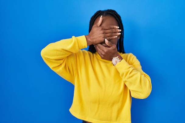 Beautiful black woman standing over blue background covering eyes and mouth with hands, surprised and shocked. hiding emotion  - Photo, Image