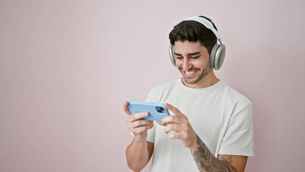 Νεαρός Ισπανόφωνος βλέποντας βίντεο στο smartphone χαμογελώντας σε απομονωμένο ροζ φόντο - Φωτογραφία, εικόνα