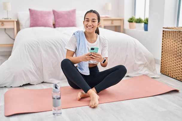 Mujer hispana joven usando teléfono inteligente sentado en el piso en el dormitorio - Foto, imagen