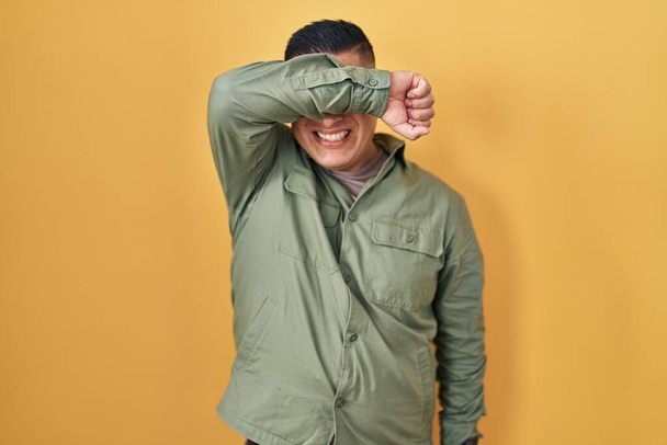 Jeune homme hispanique debout sur fond jaune couvrant les yeux avec le bras souriant gai et drôle. concept aveugle.  - Photo, image