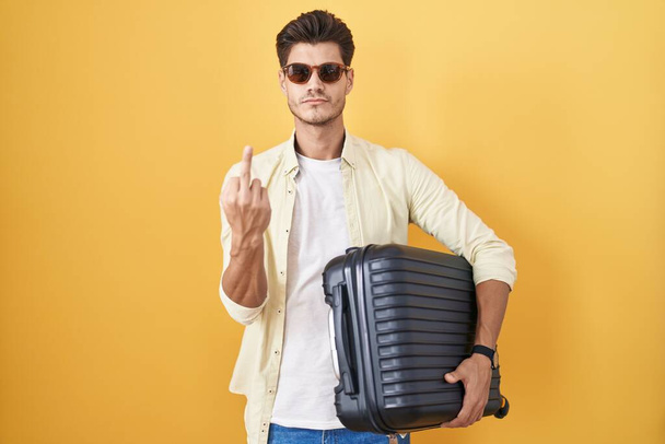 Young hispanic man holding suitcase going on summer vacation showing middle finger, impolite and rude fuck off expression  - Photo, image