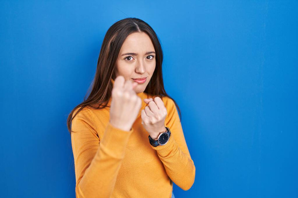 Young brunette woman standing over blue background ready to fight with fist defense gesture, angry and upset face, afraid of problem  - Zdjęcie, obraz
