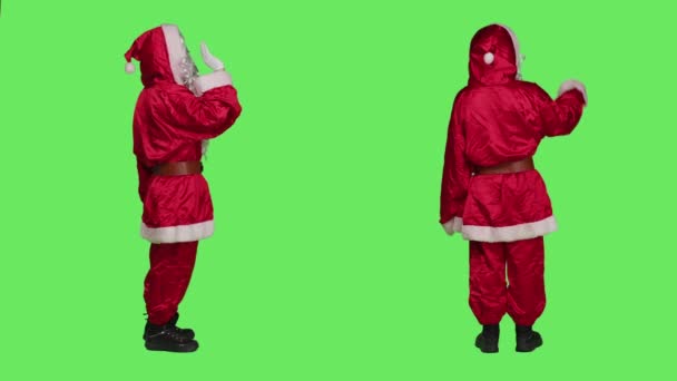 Fröhlich grüßt der Heilige Nikolaus die Menschen, die im festlich roten Anzug über dem grünen Bildschirm stehen, um saisonale Urlaubsstimmung zu verbreiten. Mann winkt Hallo und bereitet Weihnachtsfeier vor. - Filmmaterial, Video